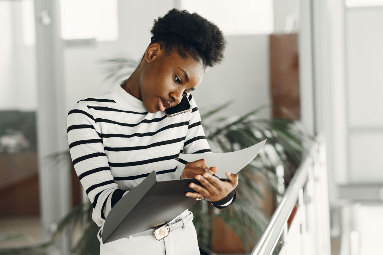 Services African American woman multitasking with phone and notepad indoors, wearing striped top.
