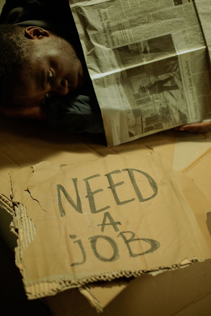 About A man lies on cardboard indoors with a 'Need a Job' sign, highlighting social issues.